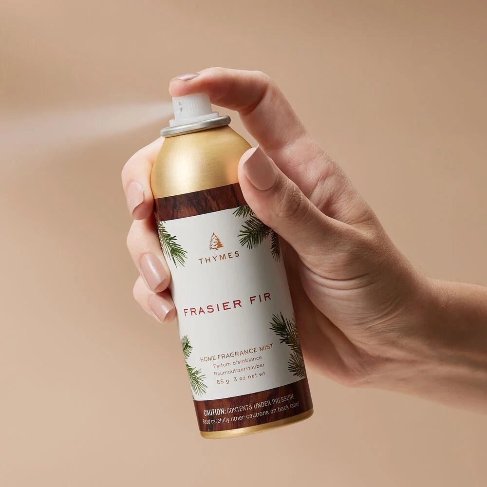 A hand holds Thymes - Frasier Fir Home Fragrance Mist 3oz, an elegant air freshener featuring a golden top and green fir branch design. The neutral beige background enhances its subtle elegance.