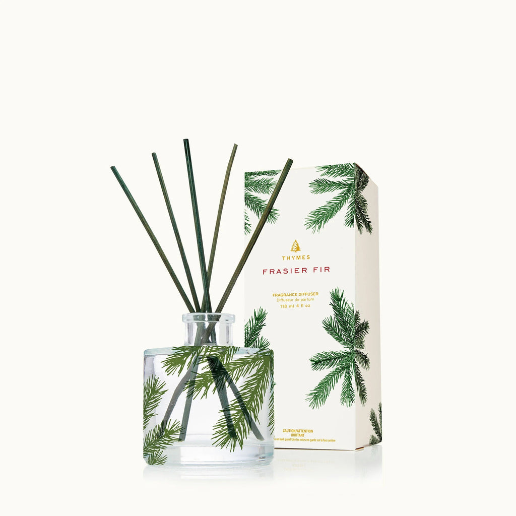 The Thymes - Frasier Fir Petite Pine Needle 4oz Reed Diffuser in a clear glass bottle with green reeds and pine branch accents exudes elegance. The white box, featuring pine branch designs and the Thymes Frasier Fir label, captures the essence of fresh pine needle aroma.