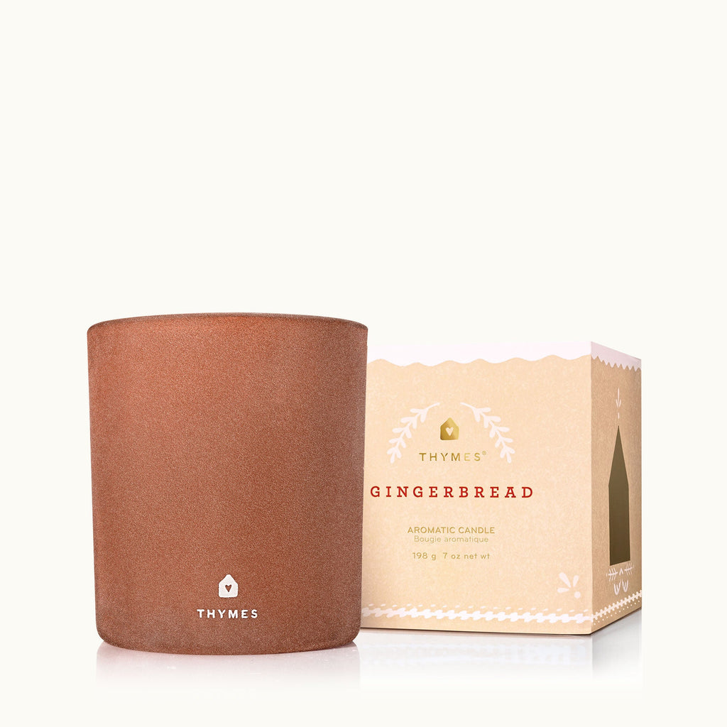 The Thymes Gingerbread 7oz Aromatic Candle, ideal for holiday baking fans, is shown beside its beige and pink box with white accents and the product name in red—a festive scent from Thymes.