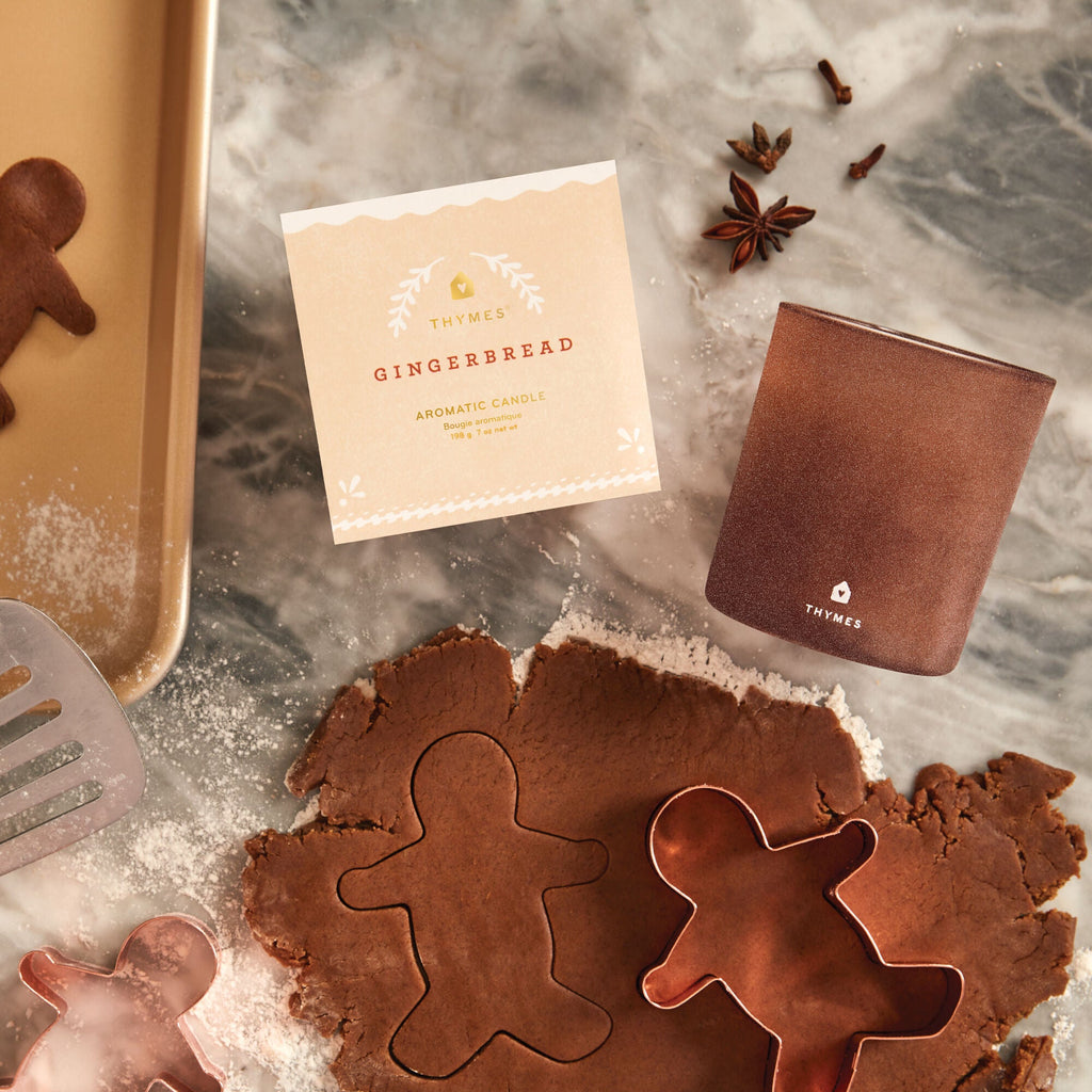 Gingerbread cookie dough and two gingerbread-shaped cookie cutters sit on a floured surface beside the Thymes - Gingerbread 7oz Aromatic Candle, with star anise and baking tools adding holiday charm to the cozy setting.
