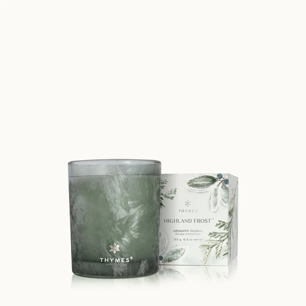 A green Thymes glass candle sits beside a botanically illustrated box labeled Thymes - Highland Frost Boxed 6.5oz Candle in sage. The soft, off-white background enhances the frosted eucalyptus theme beautifully.