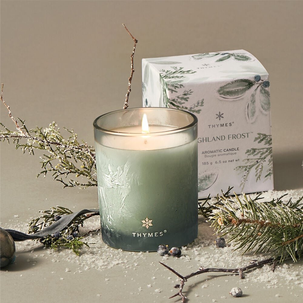 A lit Thymes - Highland Frost Boxed 6.5oz Candle labeled Thymes is next to its decorative box, surrounded by frosted eucalyptus branches, berries, and a metallic ornament on a light surface, creating a winter ambiance.