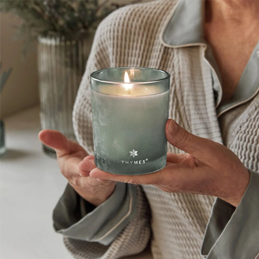 A person wearing a cozy robe holds a lit Thymes Highland Frost Boxed 6.5oz Candle in a frosted glass container. The background, echoing Faded Sage, is softly focused on greenery, creating a tranquil atmosphere.