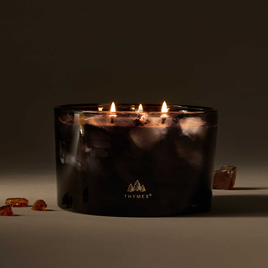 The Thymes Noir Woods 3-Wick Poured Candle, in dark marbled glass with a tree logo, glows on a neutral surface while amber stones accent the smoky wood scent and elegant design.