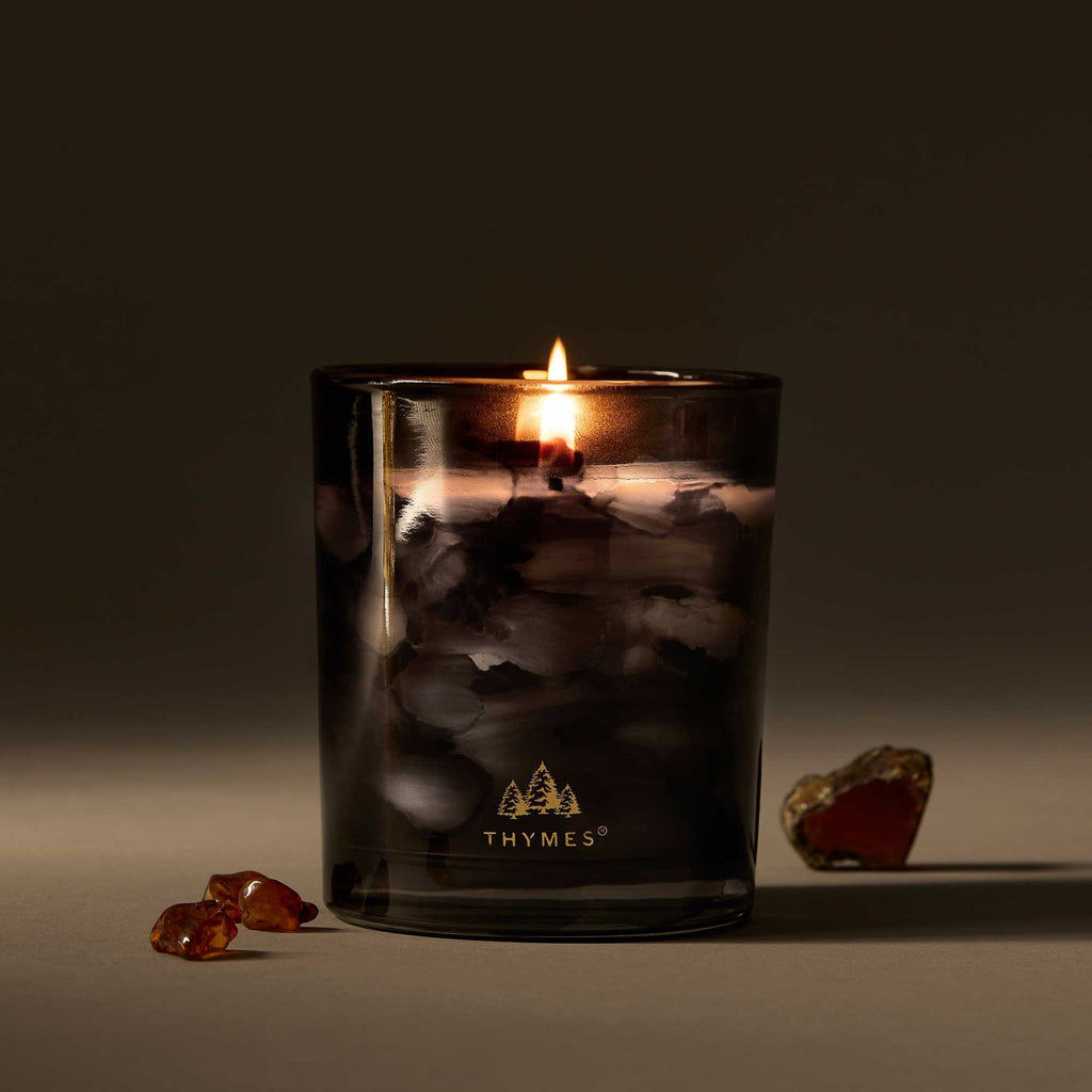 A lit Thymes Noir Woods 6.5 oz Poured Candle in a smoky glass holder with the Thymes logo sits on a surface, beside amber stones and a polished stone, against a neutral background.