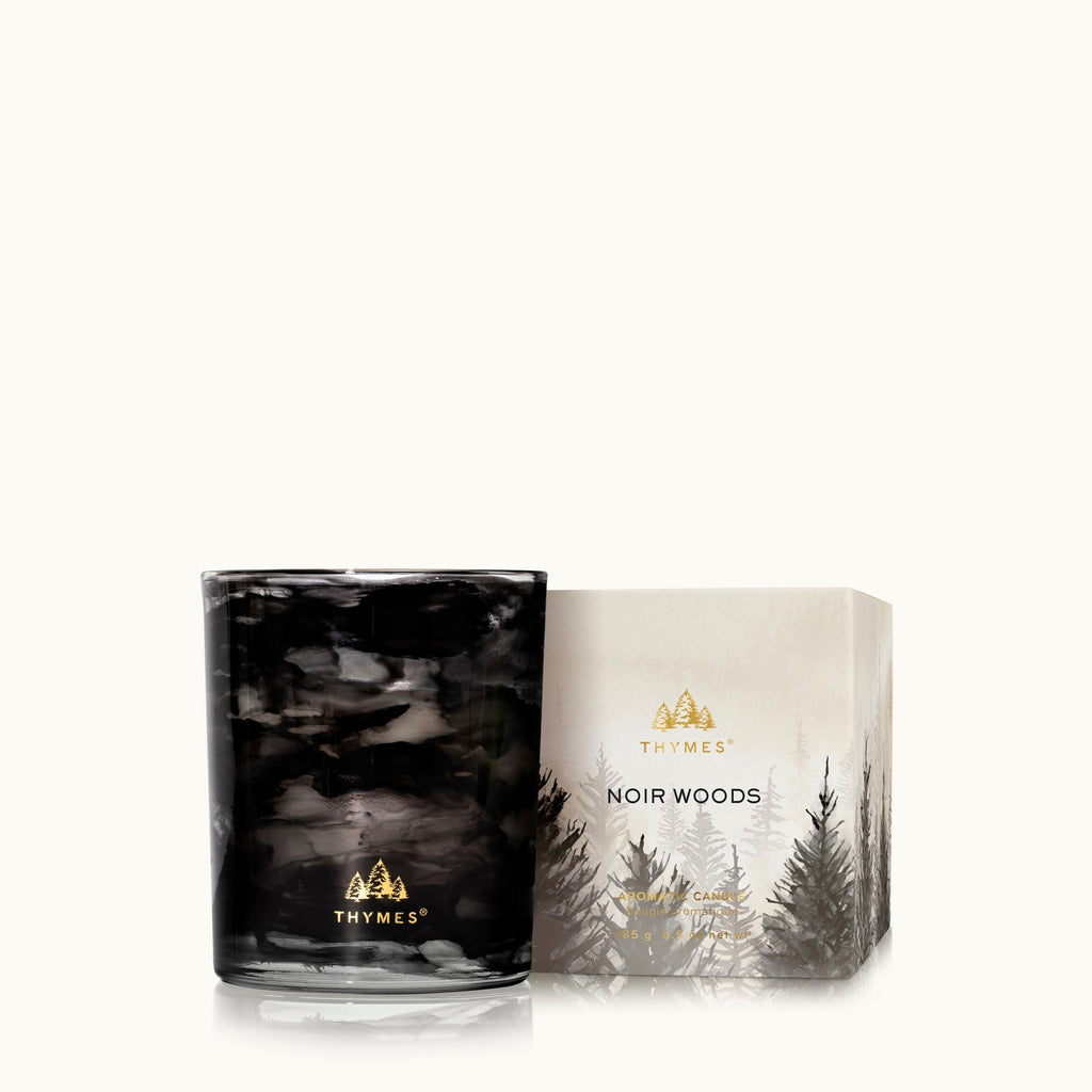 The Thymes Noir Woods 6.5 oz Poured Candle, in black marbled glass with a gold logo, is shown beside its decorative box featuring a soft gray and gold forest design.