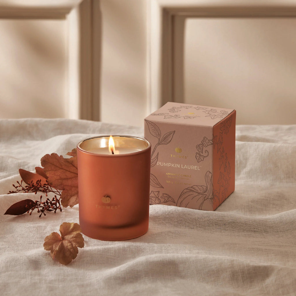 The Thymes Pumpkin Laurel 6.5oz Aromatic Candle rests on fabric beside dried autumn leaves and its matching box, filling the cozy space with inviting pumpkin fragrance.