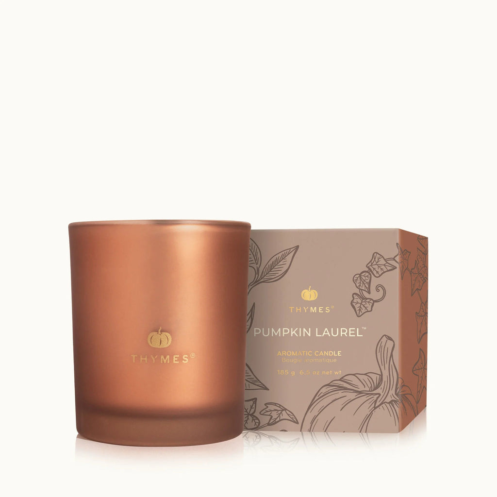 The Thymes Pumpkin Laurel 6.5oz Aromatic Candle, in a copper-colored jar, sits beside its decorative box with botanical artwork—an ideal way to add a warm autumn scent to your home.