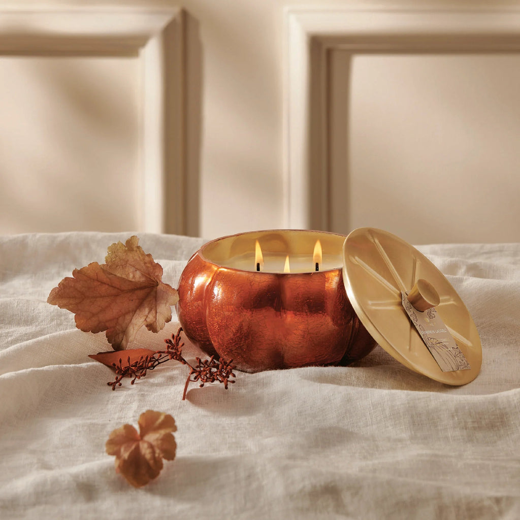 The Thymes Pumpkin Laurel Pumpkin Shape 15oz Aromatic Candle burns with three flames on a white cloth, accompanied by a beige lid, matches, and autumn leaves—creating the perfect cozy fall decor.