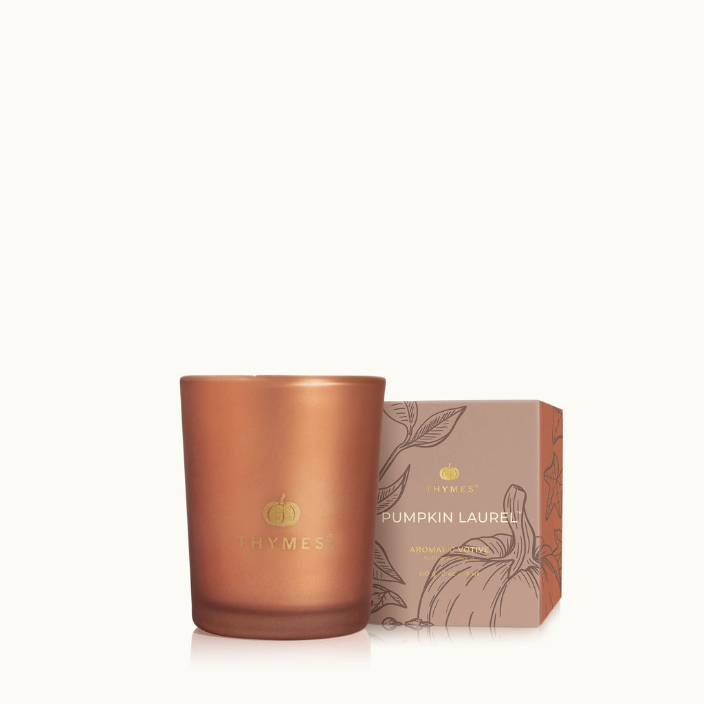 The Thymes - Pumpkin Laurel Votive Aromatic Candle sits beside its illustrated, gold-accented box. This copper-colored candle from Thymes makes an ideal autumn hostess gift, beautifully displayed on a light background.
