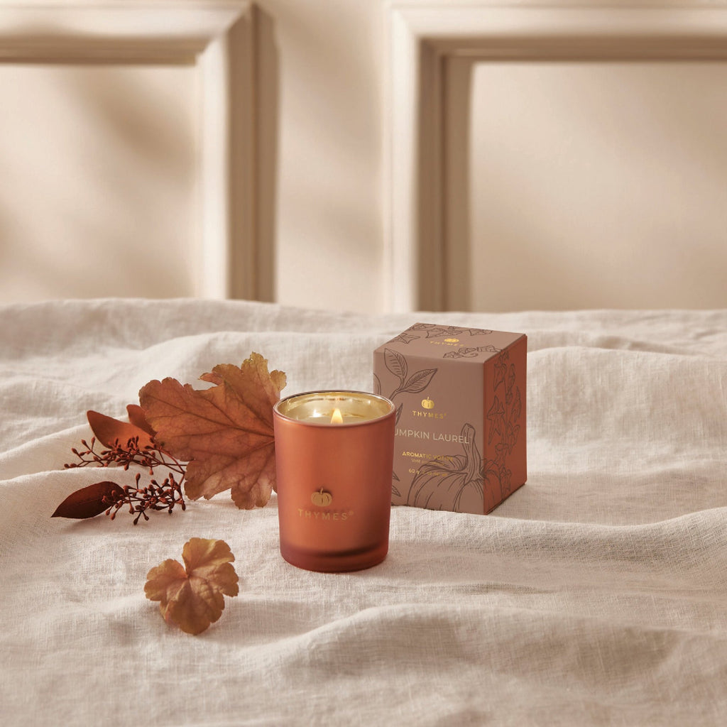 The Thymes Pumpkin Laurel Votive Aromatic Candle in a glass holder sits beside autumn leaves and a matching box on a white cloth. Its cozy fall scent makes it the perfect hostess gift for any room with elegant neutral decor.