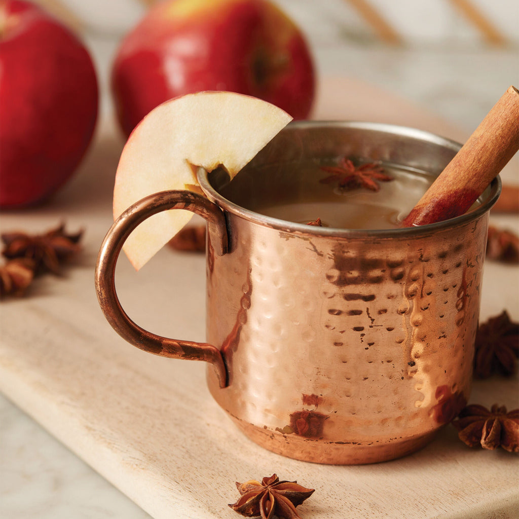 A Thymes Shimmered Cider Copper Mug 10oz Aromatic Candle evokes cozy cider aromas, styled with a cinnamon stick, star anise, and apple slice on a wooden board. Red apples and more star anise appear in the background.