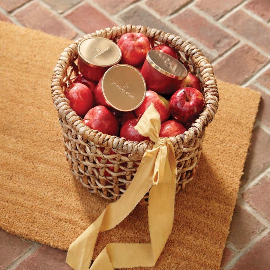 A woven basket with a yellow ribbon holds red apples and Thymes - Simmered Cider Candle Tins with gold lids, set on a textured brown doormat over brick.