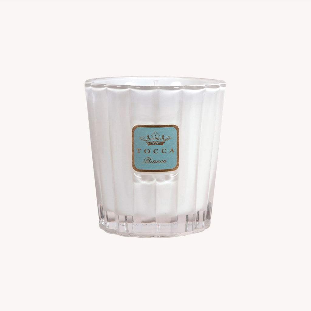 A white luxury candle in a ribbed glass holder with a blue and gold label reading Tocca - Bianca 10oz Candle features a subtle green tea fragrance set against a plain, light backdrop.