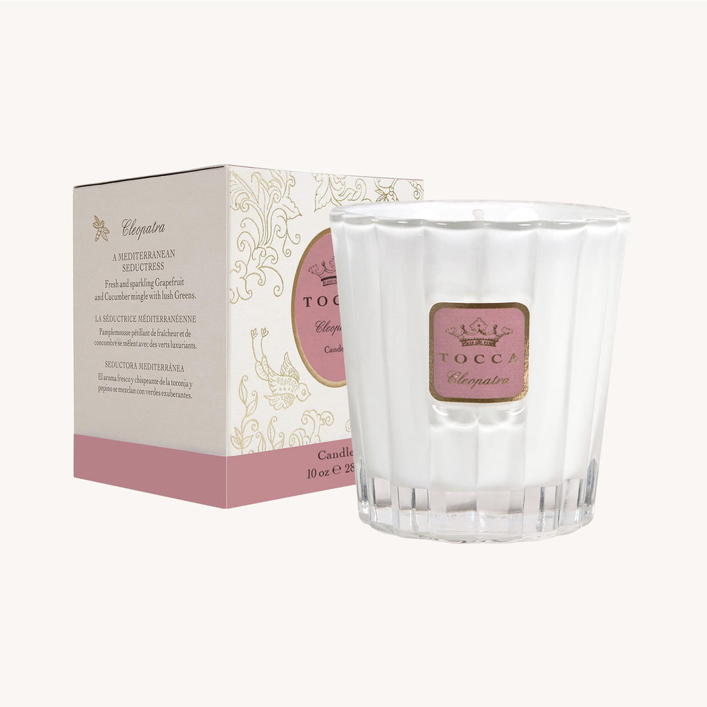 A white 10oz Tocca Cleopatra candle with a pink label sits elegantly beside its matching box, both showcasing intricate pink and gold designs. The luxurious soy-wax candle is cruelty-free.