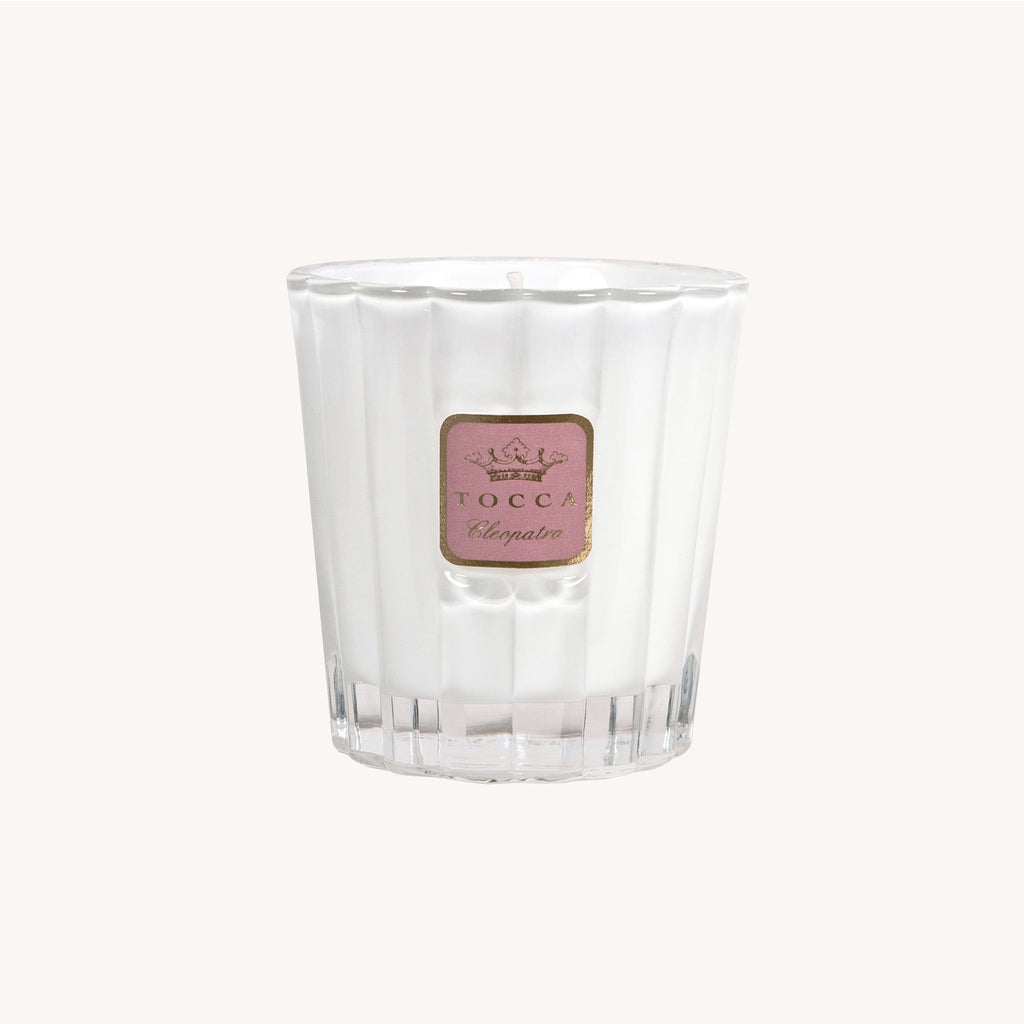 An unlit Tocca - Cleopatra 10oz luxury soy-wax candle, featuring a pink labeled fluted glass holder with the brand name Tocca and Cleopatra, is placed elegantly against a plain, light-colored background. Cruelty-free.