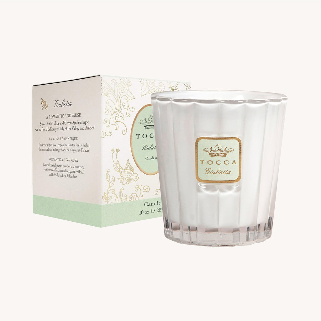 A luxury Tocca candle (10oz) in a clear glass holder labeled Giulietta sits beside its decorative pale green and white box, featuring elegant designs and detailing the scent with notes of green apple and pink tulips.