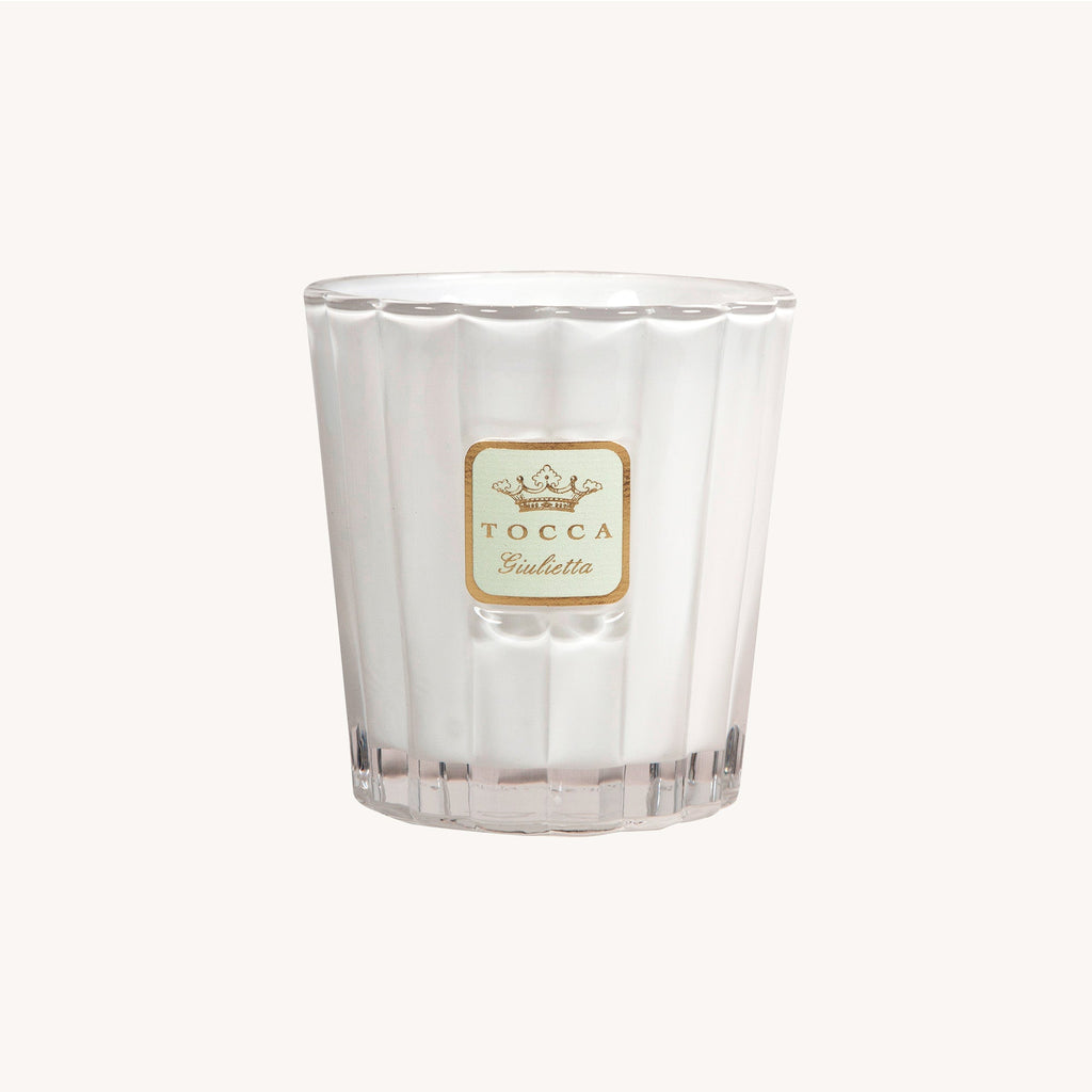 The Tocca - Giulietta 10oz Candle, in a ribbed jar with a gold Tocca Giulietta label, elegantly sits on a plain backdrop. Its unlit fragrance of fresh green apples invites an inviting atmosphere to any space.