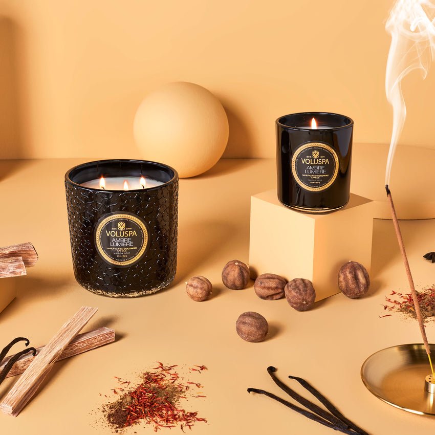 Voluspas Ambre Lumiere Classic Candle in black jars sits on a beige surface surrounded by incense, wooden sticks, vanilla pods, nuts, coconut wax elements, and saffron strands. A spherical vase and cubes add decoration as clean-burning smoke wafts gently from the burning incense.