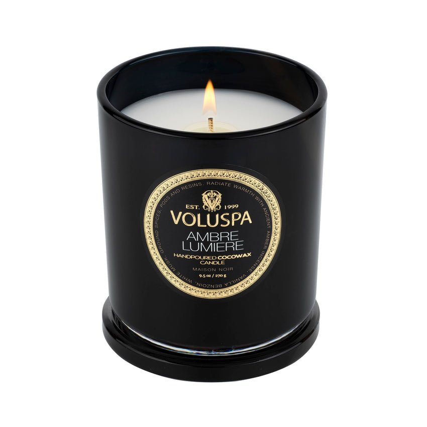 The Voluspa - Ambre Lumiere Classic Candle 9.5oz, made with clean-burning coconut wax, sits in a black glass holder with a golden label. Its white wax melts atop the matching black lid, releasing amber incense notes into the air.