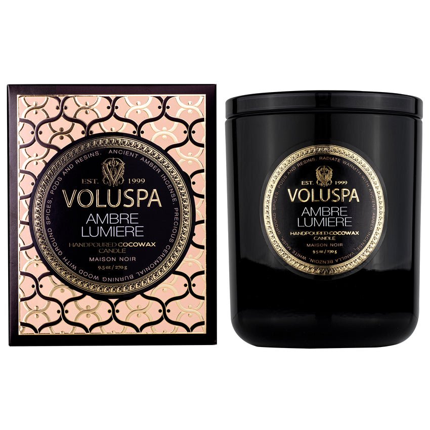 The Voluspa - Ambre Lumiere Classic Candle 9.5oz features a sophisticated black container with gold text, packaged in an intricate gold and black box. Crafted from clean-burning coconut wax, the packaging highlights Voluspas elegant design and luxurious appeal.