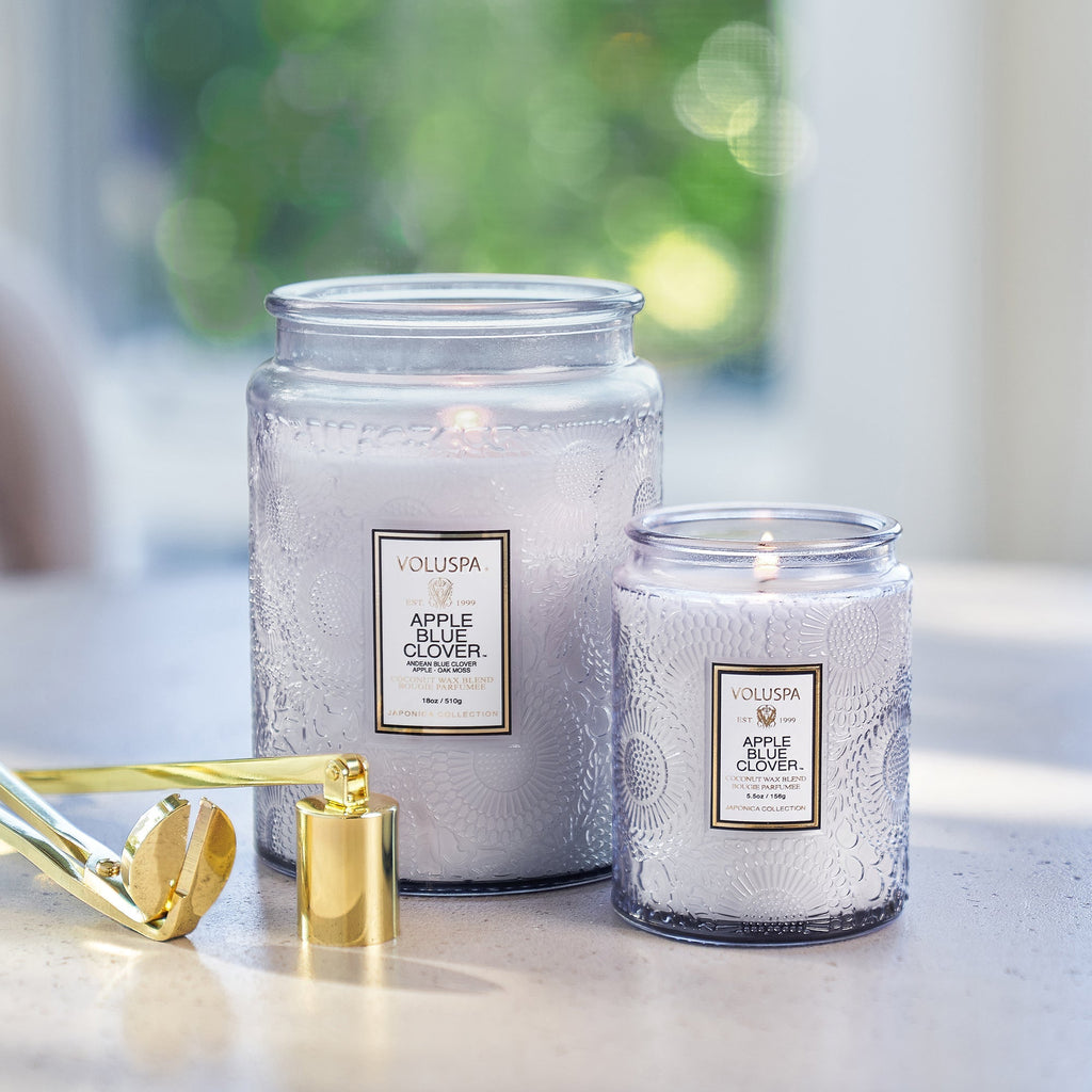 Two lit Voluspa candles labeled Apple Blue Clover on the table, crafted with coconut wax. One large in textured glass jars. A wick trimmer and candle snuffer nearby as natural light enhances their fragrance throw.