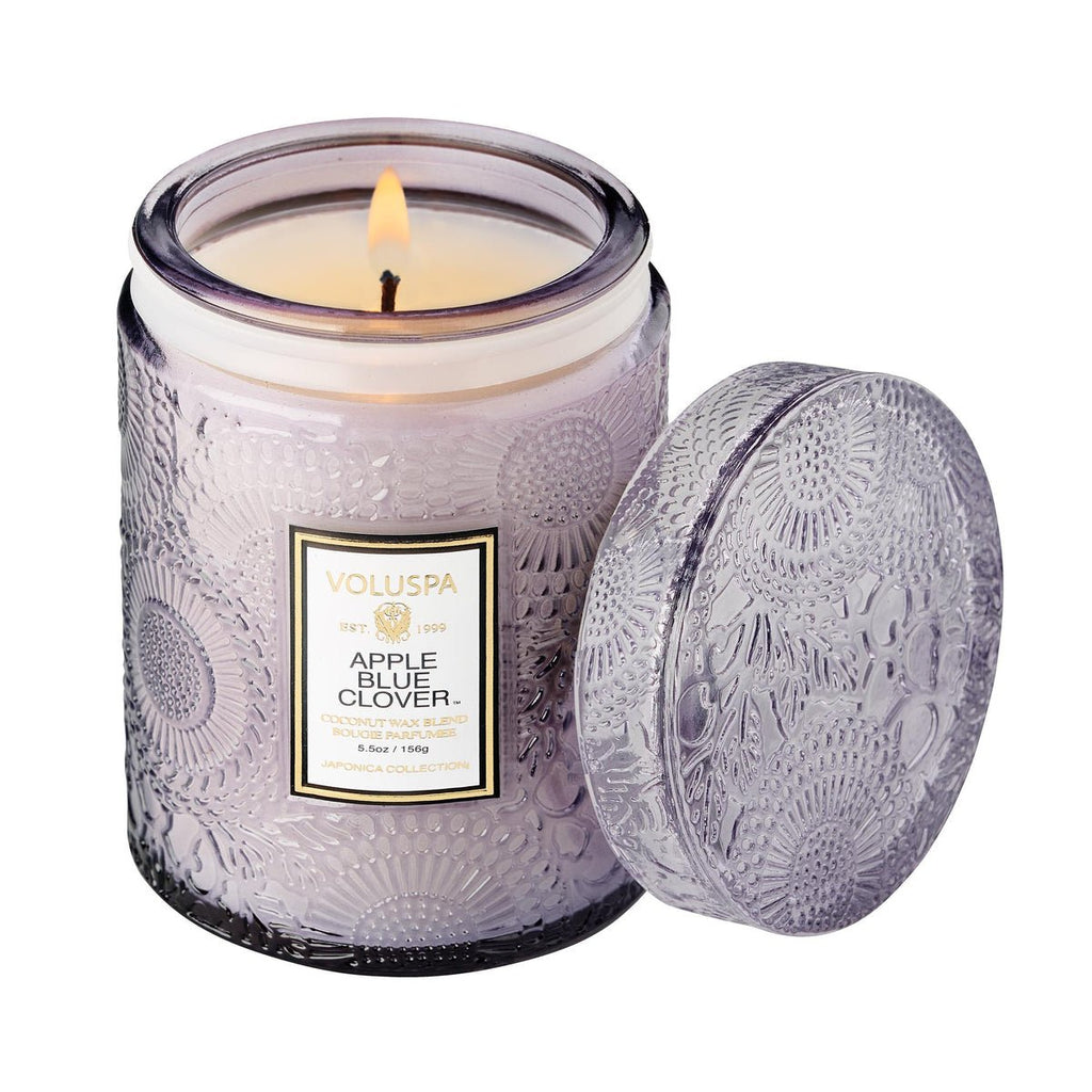 A Voluspa - Apple Blue Clover Small Jar 5.5oz Candle is lit, with the lid set aside. It features a coconut wax blend for impressive fragrance throw, while the jar and lid display an intricate embossed pattern.