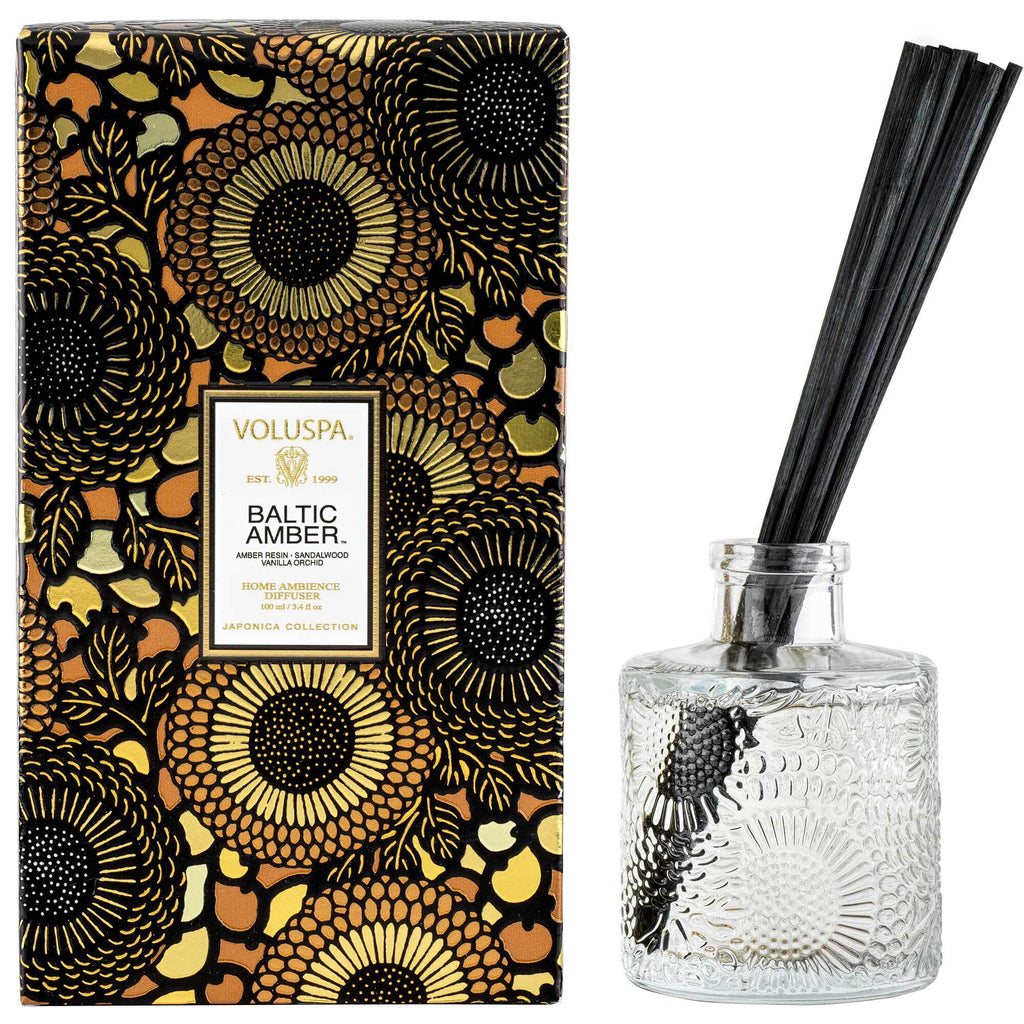 A Voluspa Baltic Amber 3.4oz Reed Diffuser features a floral-patterned gold and black box beside a glass diffuser bottle embossed with floral motifs. Five black reeds extend from the transparent bottle, creating a charming display.