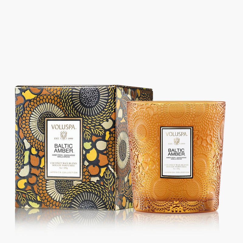 The Voluspa - Baltic Amber Classic 9oz Candle rests in an embossed amber glass jar next to its floral-patterned box with gold, black, and orange accents, elegantly arranged on a white surface.