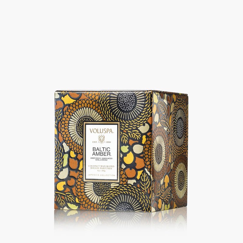The Voluspa Baltic Amber Classic 9oz Candle features a floral and geometric design in gold, black, and amber tones, with a front label displaying the Voluspa brand and Baltic Amber scent details.