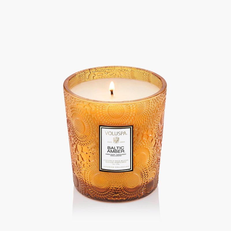 The Voluspa - Baltic Amber Classic 9oz Candle features amber coconut wax in an ornate textured glass jar, shown lit with a single flame against a plain white background.