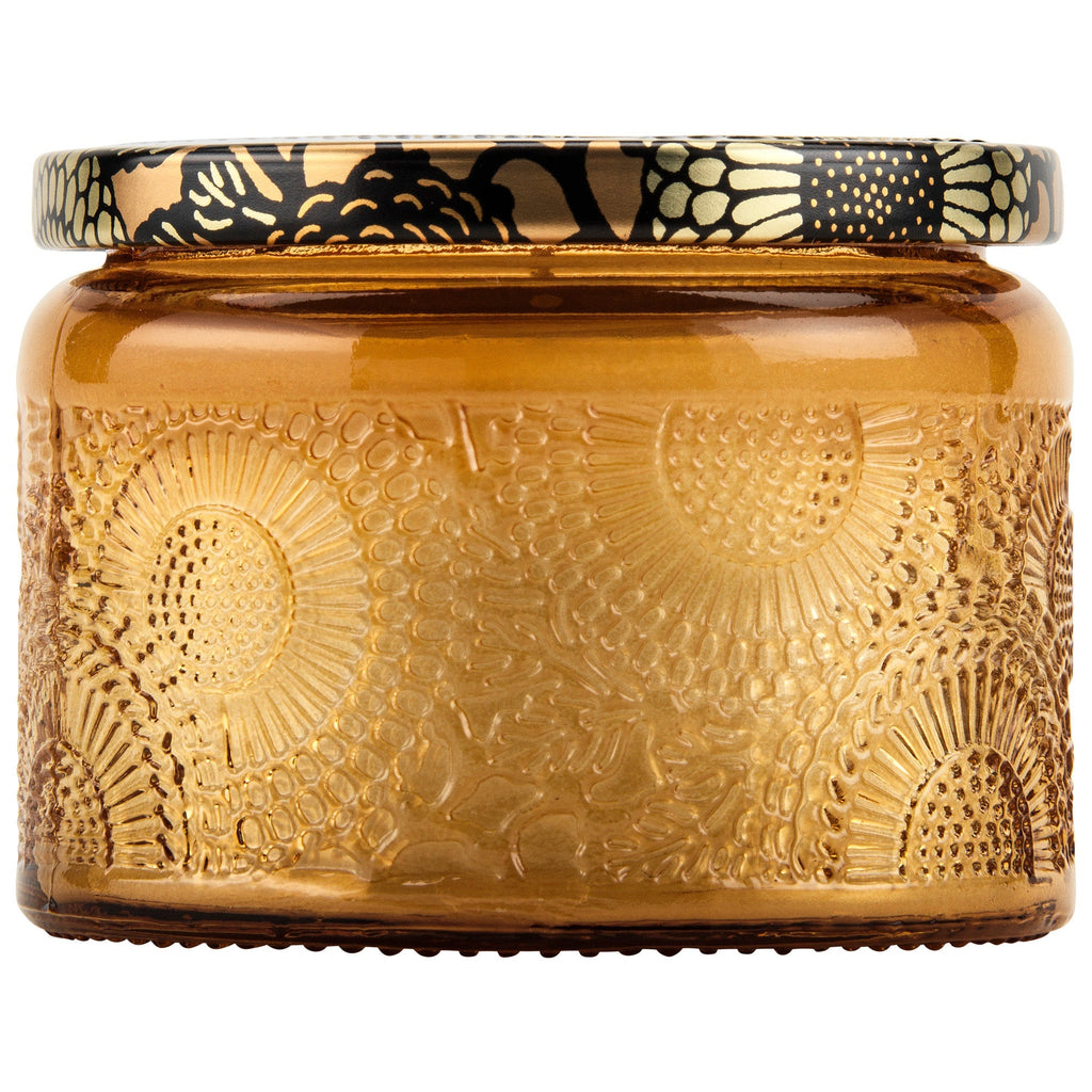 The Voluspa Baltic Amber Petite 3.2oz Jar Candle features a decorative amber glass jar with an embossed floral pattern and a gold-and-black metal lid. Made from a Baltic Amber coconut wax blend, it exudes elegance and a warm, inviting aroma.