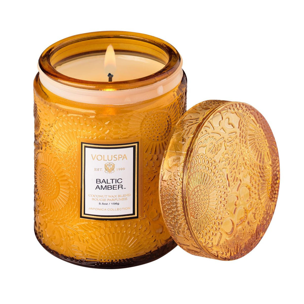 A Voluspa Baltic Amber Small Jar Candle sits beside it, crafted from coconut wax in an embossed amber glass jar with a matching lid. Its rich amber resin scent fills the air.