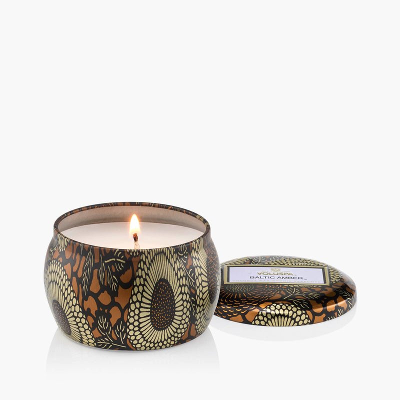 The Voluspa - Baltic Amber Tin 4oz Candle features a black, gold, and olive floral design. Crafted with a coconut wax blend, it includes a matching lid with a white label and gold trim resting beside the lit candle.