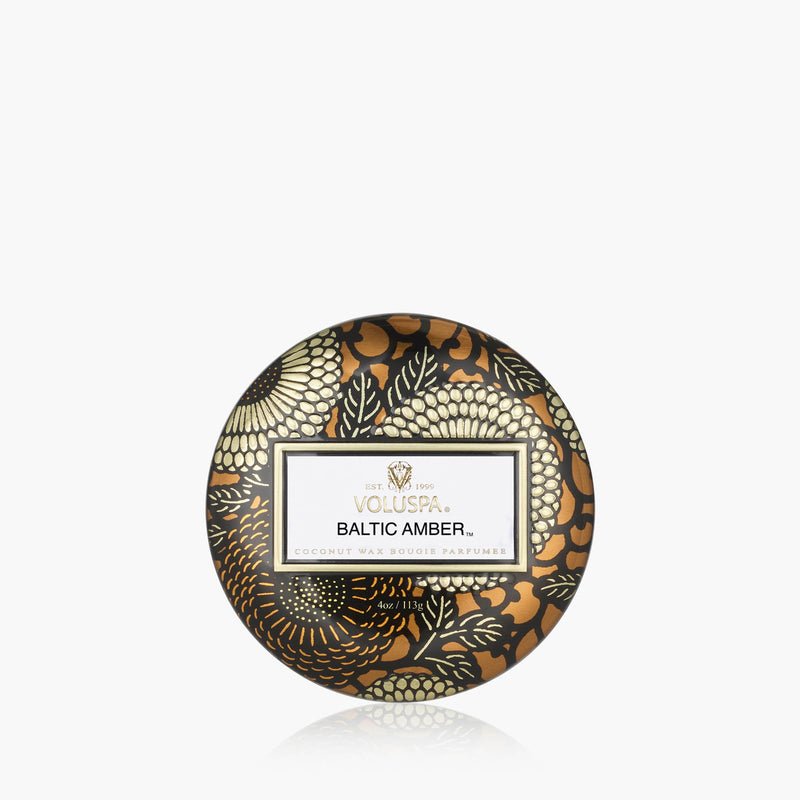 The Voluspa - Baltic Amber Tin 4oz Candle features a luxurious coconut wax blend in a round mini tin with a black, gold, and cream floral pattern and a central label on the lid.