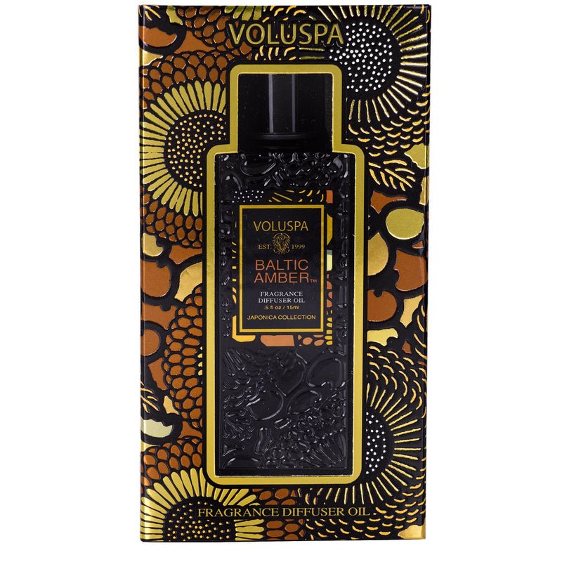 A Voluspa Baltic Amber Ultrasonic Diffuser Fragrance Oil package features a black and gold box with floral patterns, showcasing an ornate Vanilla Orchid-infused bottle through a transparent window. The label says Japonica Collection.