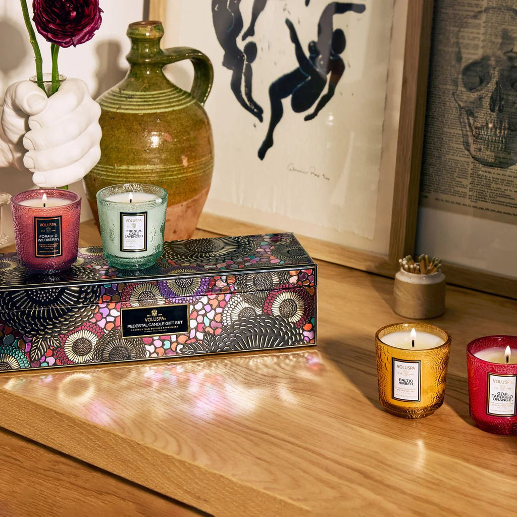 A cozy room features a Voluspa Best Sellers Pedestal Candle made of coconut wax blend, casting a delightful fragrance. Three lit colorful candles, a ceramic vase, framed art adorn the table. A patterned gift box and hand-shaped vase complete the inviting ambiance.