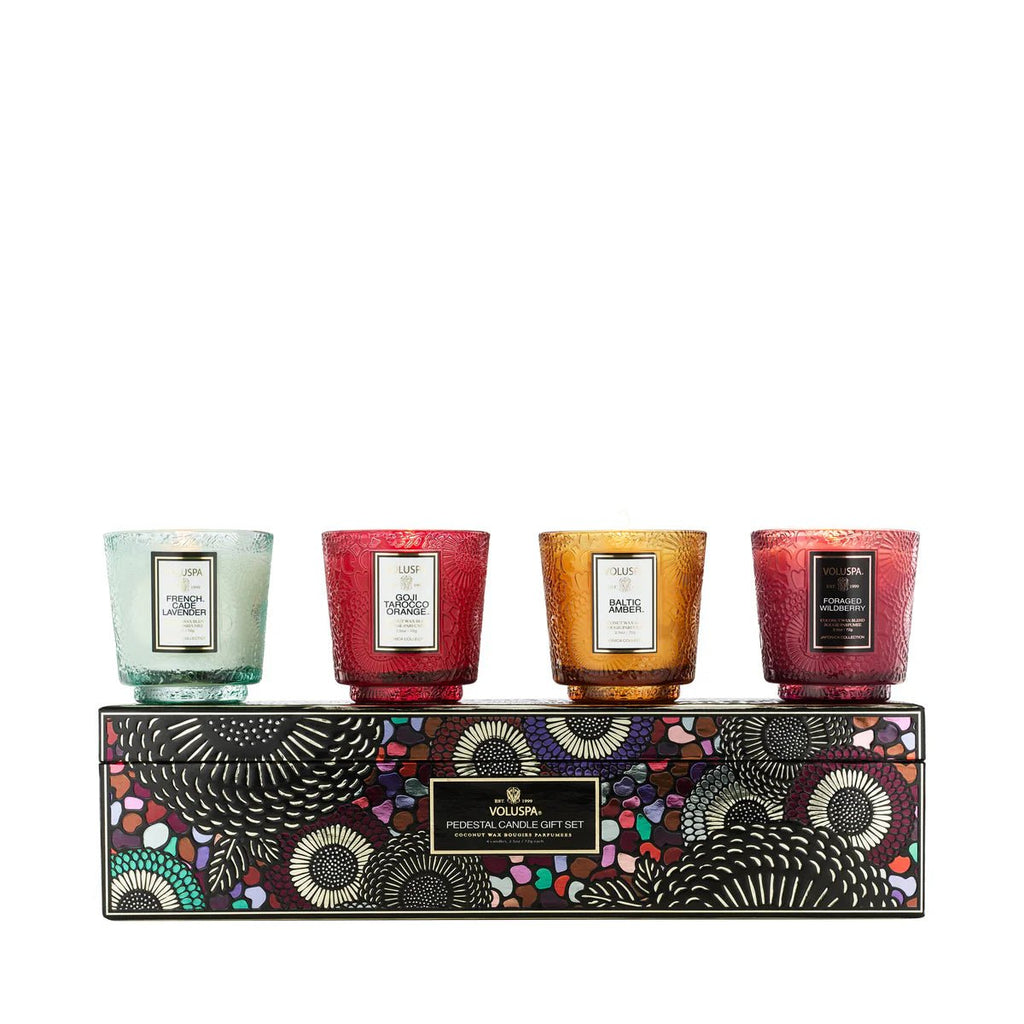 The Voluspa - Best Sellers Pedestal Candle Gift Set features four petite coconut wax candles in colorful glass holders on a patterned box. Each label highlights the impressive fragrance throw, with the box showcasing bold, abstract designs in vibrant hues.