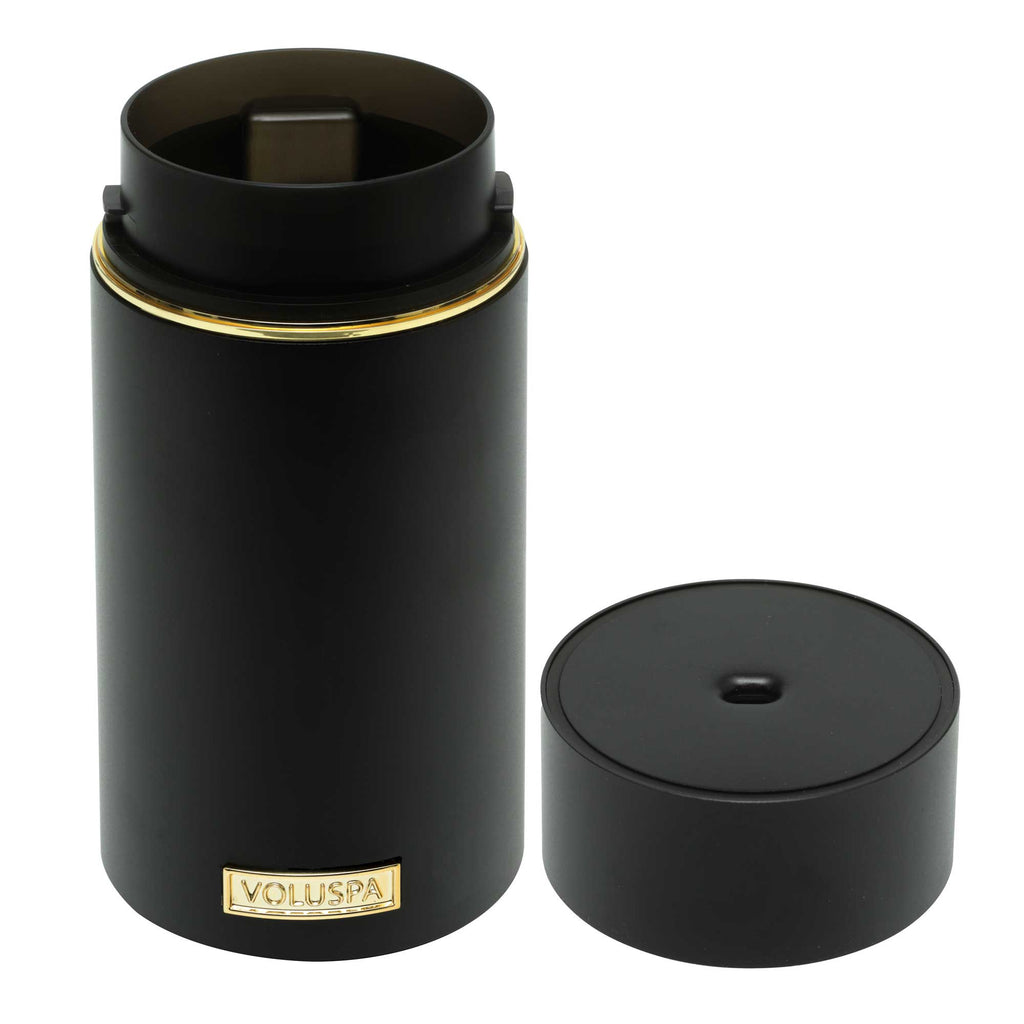 The Voluspa Black Ultrasonic Fragrance Oil Diffuser Device, with its sleek black cylindrical design and elegant gold details, is reminiscent of a luxurious candle container. The label features embossed gold lettering, adding to its sophisticated appeal.