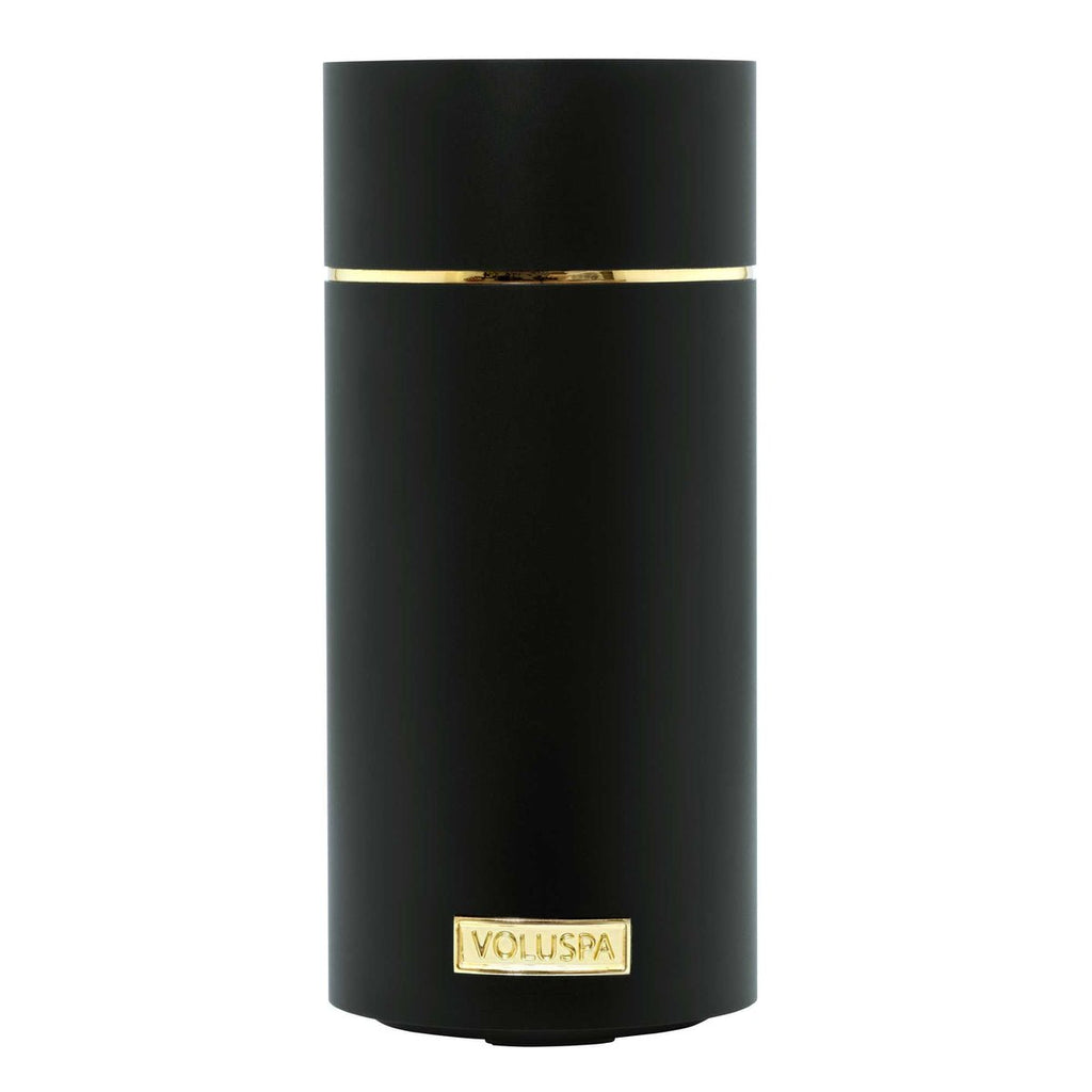 The Voluspa Black Ultrasonic Fragrance Oil Diffuser Device has a cylindrical design with a sleek black finish, complemented by a gold band near the top and a gold label at the bottom center.