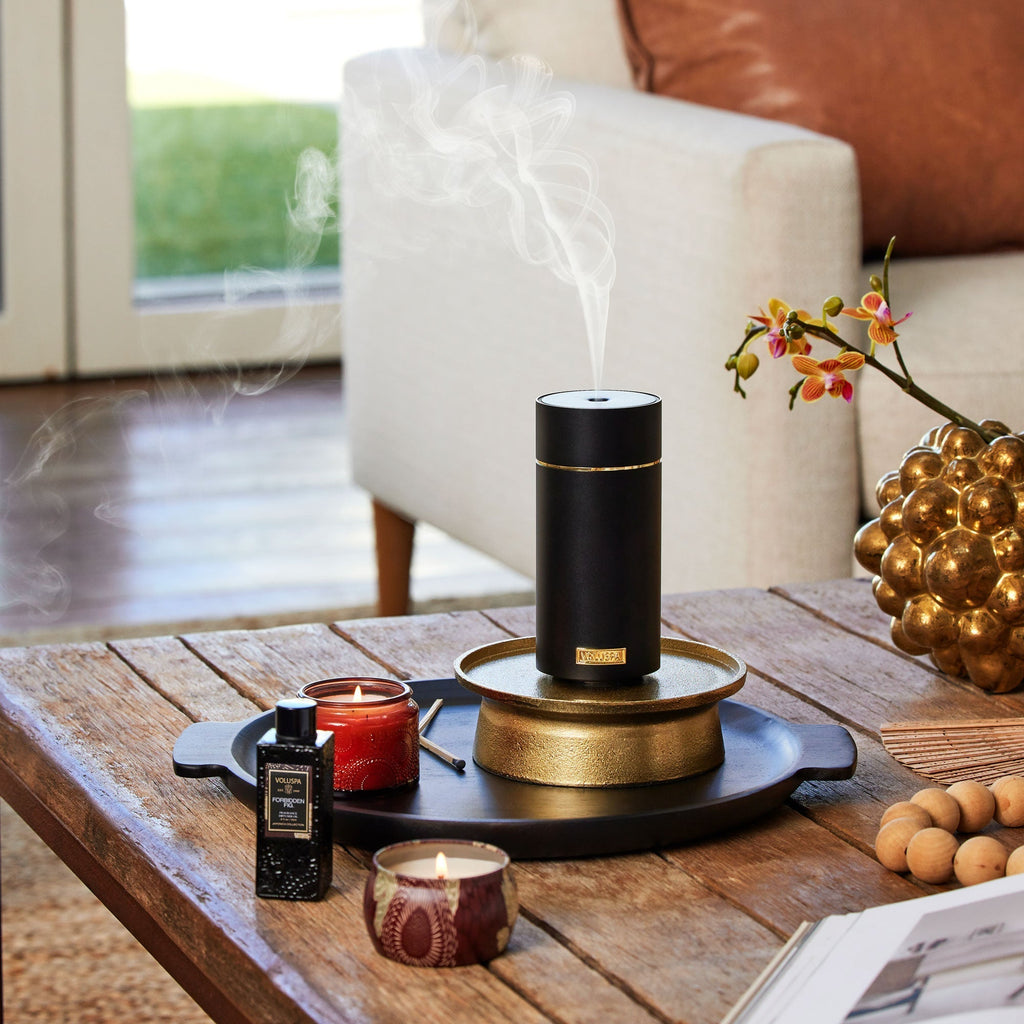 A cozy living room displays a wooden coffee table with the Voluspa Black Ultrasonic Fragrance Oil Diffuser emitting mist, several lit candles, a decorative gold orb, and a small bottle. In the background, a white couch with a leather pillow enhances the inviting ambiance.