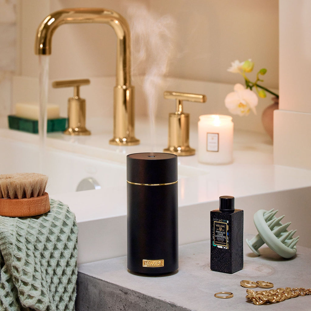 A bathroom countertop features a Voluspa - Black Ultrasonic Fragrance Oil Diffuser emitting steam beside a textured black bottle. A lit candle and orchid enhance the ambiance near a gold faucet, while a brush, green cloth, hair tool, and gold jewelry complete the serene setting.