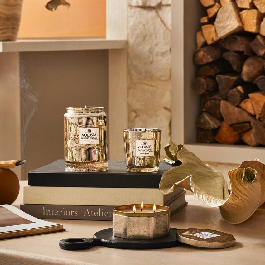 A cozy room showcases a lit Voluspa - Blond Tabac Large Jar 18oz Candle on the coffee table with books. The coconut wax blend offers a comforting scent, as the decorative leaf and stacked firewood in the background enhance the warm ambiance.