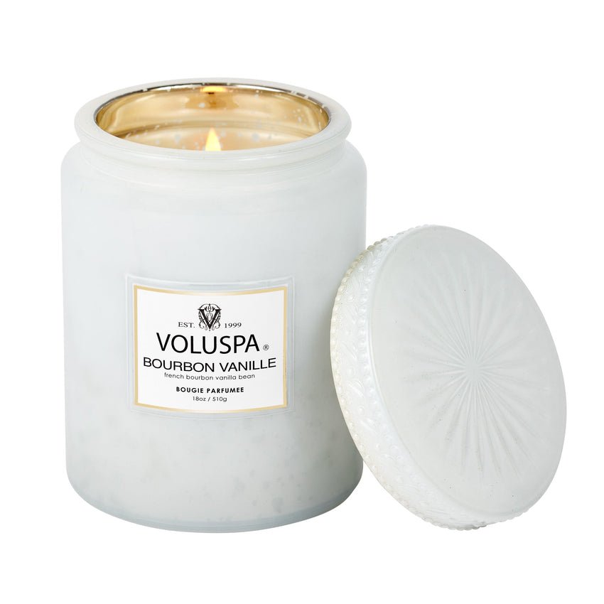 A lit Voluspa Bourbon Vanille 18oz candle, made with a silky coconut wax blend, sits in a frosted glass jar. Its textured lid leans against it, while the elegant label displays the brand and scent of this exquisite gourmand fragrance.