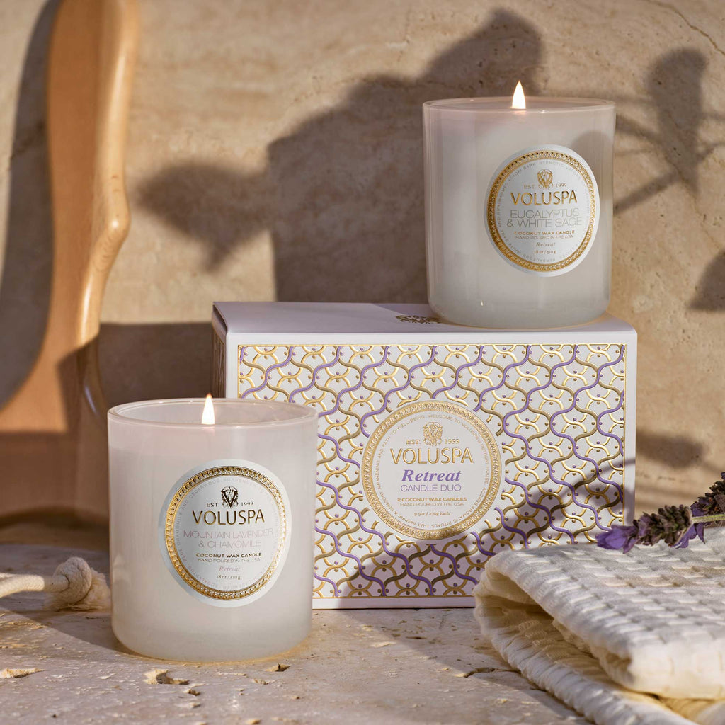 Two lit Voluspa candles from the California Summers Getaway Duo are displayed with a decorative box, wooden brush, white towel, and lavender sprigs on a beige surface.