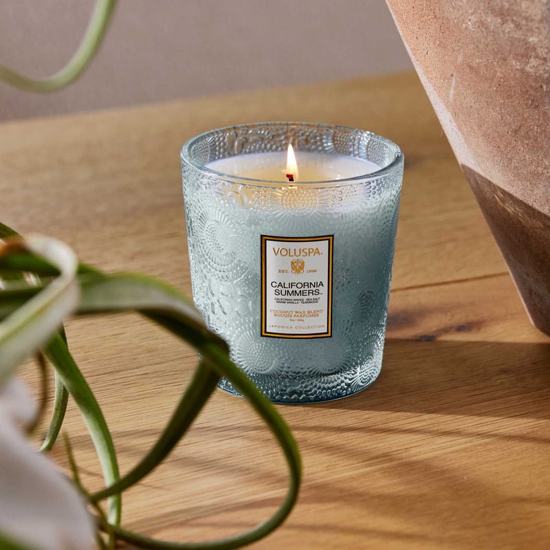 A Voluspa California Summers Getaway Duo Gift Set candle glows in a textured blue jar on a wooden surface, next to a terracotta pot and green and white plant leaves.