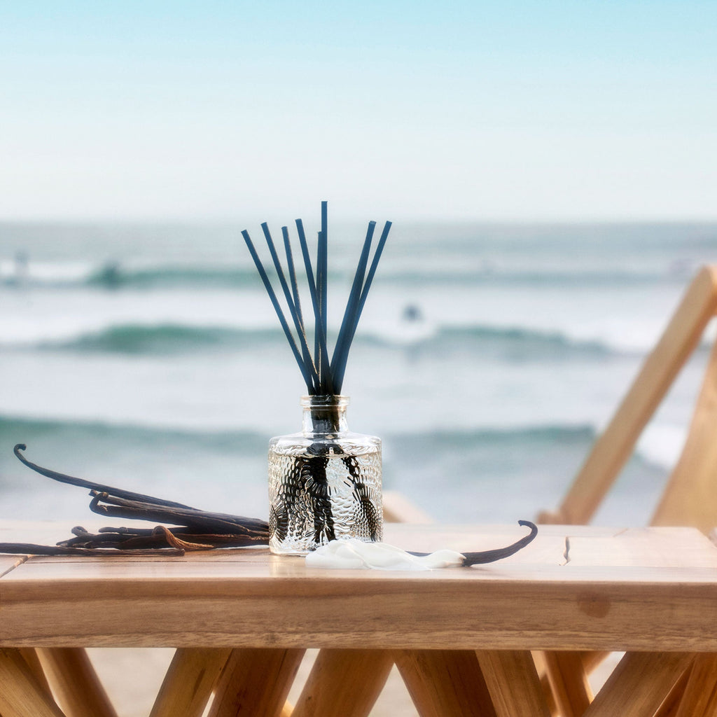 The Voluspa California Summers Getaway Duo Gift Set sits on a wooden table by the beach, with black diffuser sticks, vanilla beans, and a seashell. In the background, waves roll in and beach chairs line the shore.