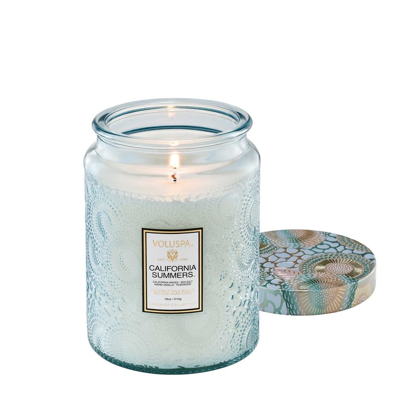 A Voluspa California Summers candle in a large 18oz textured glass jar emits a warm glow. Crafted from a coconut wax blend in a subtly blue-tinted glass, it features an intricately patterned floral lid.