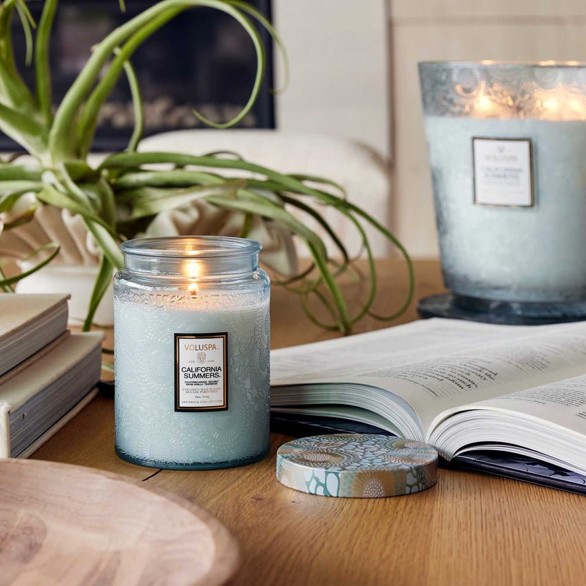 A Voluspa - California Summers Large Jar 18oz Candle made of a coconut wax blend sits on a wooden table, its aroma filling the air. Nearby lie an open book, a small wooden bowl, and a decorative plant with long green leaves.