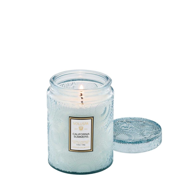 A lit Voluspa - California Summers Small Jar Candle in a decorative clear glass jar with an embossed lid beside it features the label Voluspa California Summers. Made from a coconut wax blend, this clean-burning 5.5oz candle emits fresh aquatic notes, with slightly melted wax by the flame.